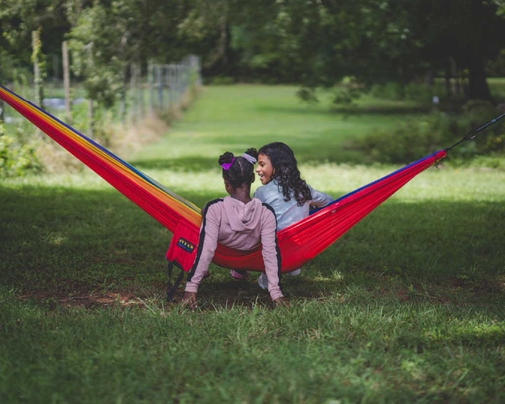 two Black children sitting in a hammock facing away from the camera. The scenery is lush green grass and trees in a land sanctuary in Durham, NC. 