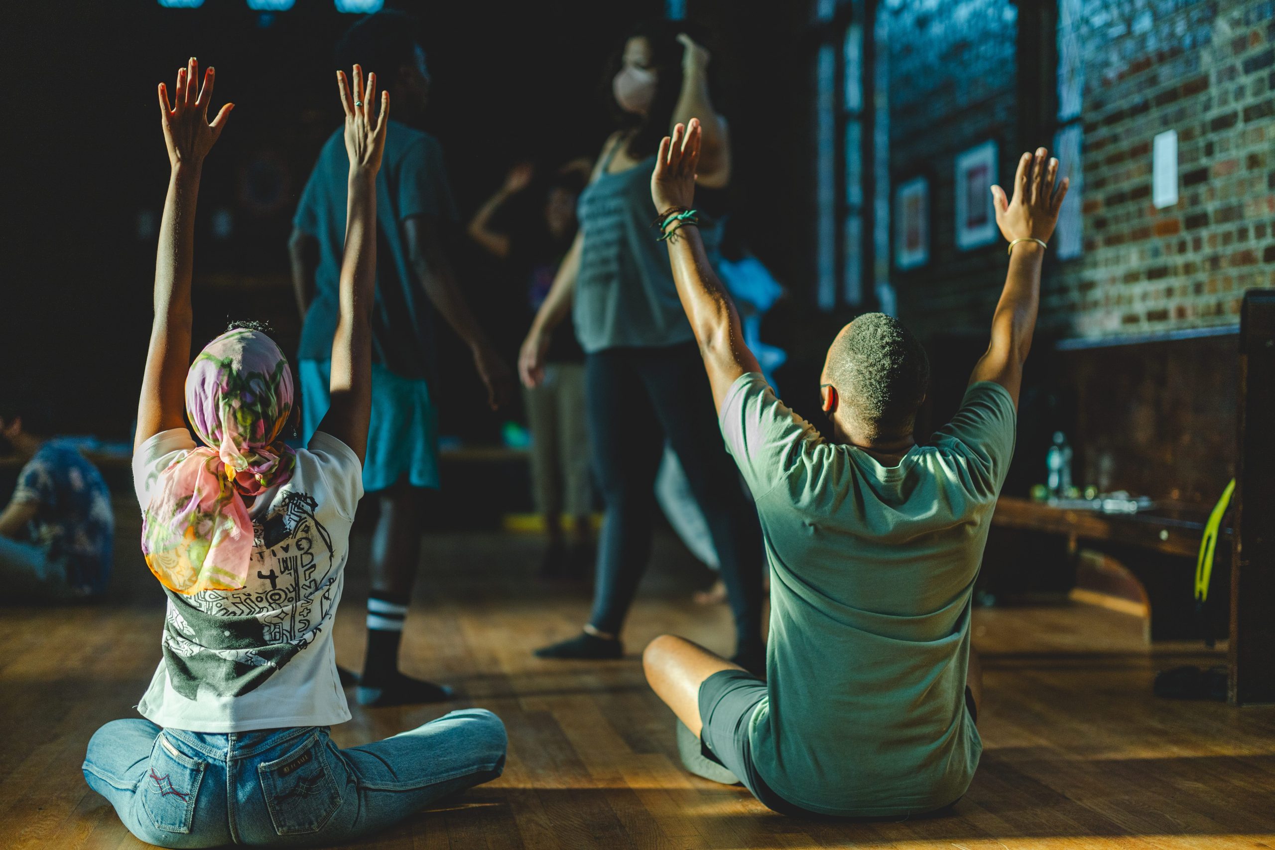 BIPOC community members participating in a somatic body workshop. Two people are sitting on the floor, backs to the camera, holding up their arms to the sky. Several other people are standing and talking behind them. They are in a church with natural lighting coming from the left.