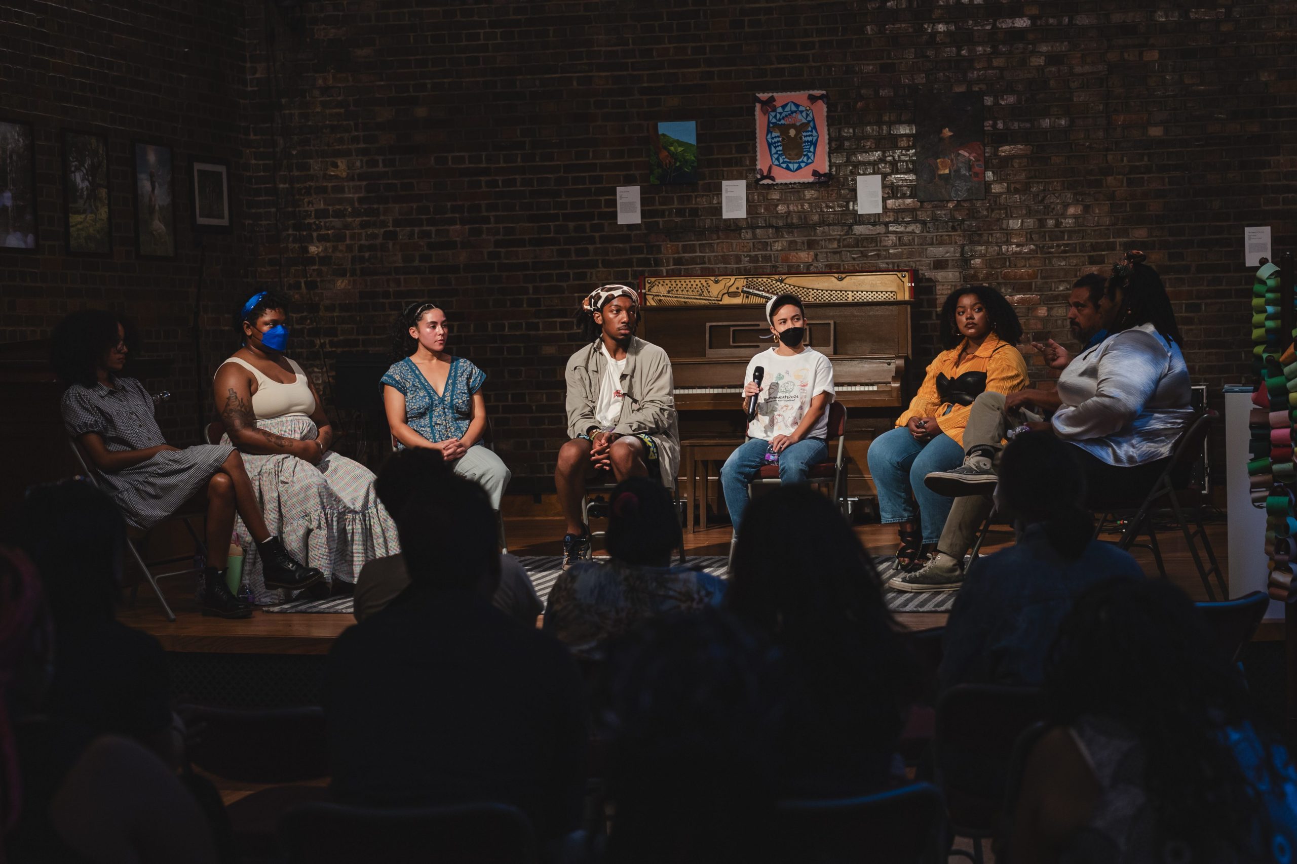 Six BIPOC artists are seating in a semi-circle on a stage in a church. They are speaking on an artist panel. Artwork on panels surround them behind them. The room is dimly lit with a spotlight on the artists on stage.
